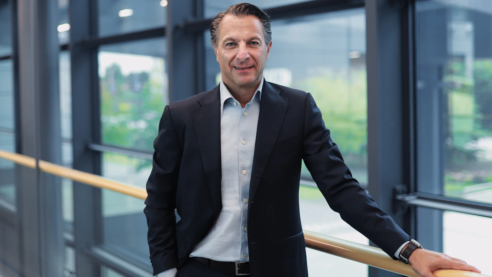 A man in a dark suit and light dress shirt stands by a handrail indoors, tesa tape nearby, large windows behind him. He smiles gently. (This text has been generated by AI)
