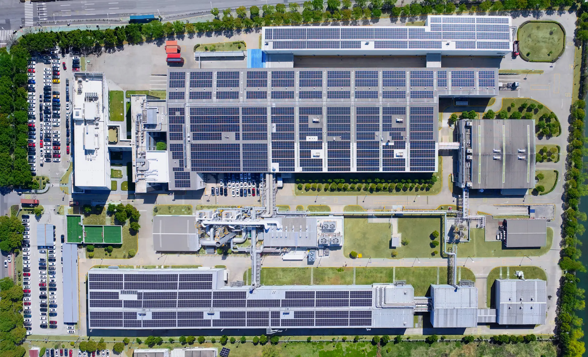 Aerial view of an industrial facility with many buildings, tesa tape banners on rooftops with solar panels, lawns, trees, and parked cars.