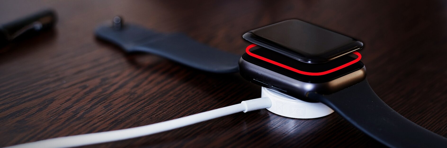 A black smartwatch with a black wristband rests on a white wireless charging dock, secured with tesa tape, on a dark wooden surface.
