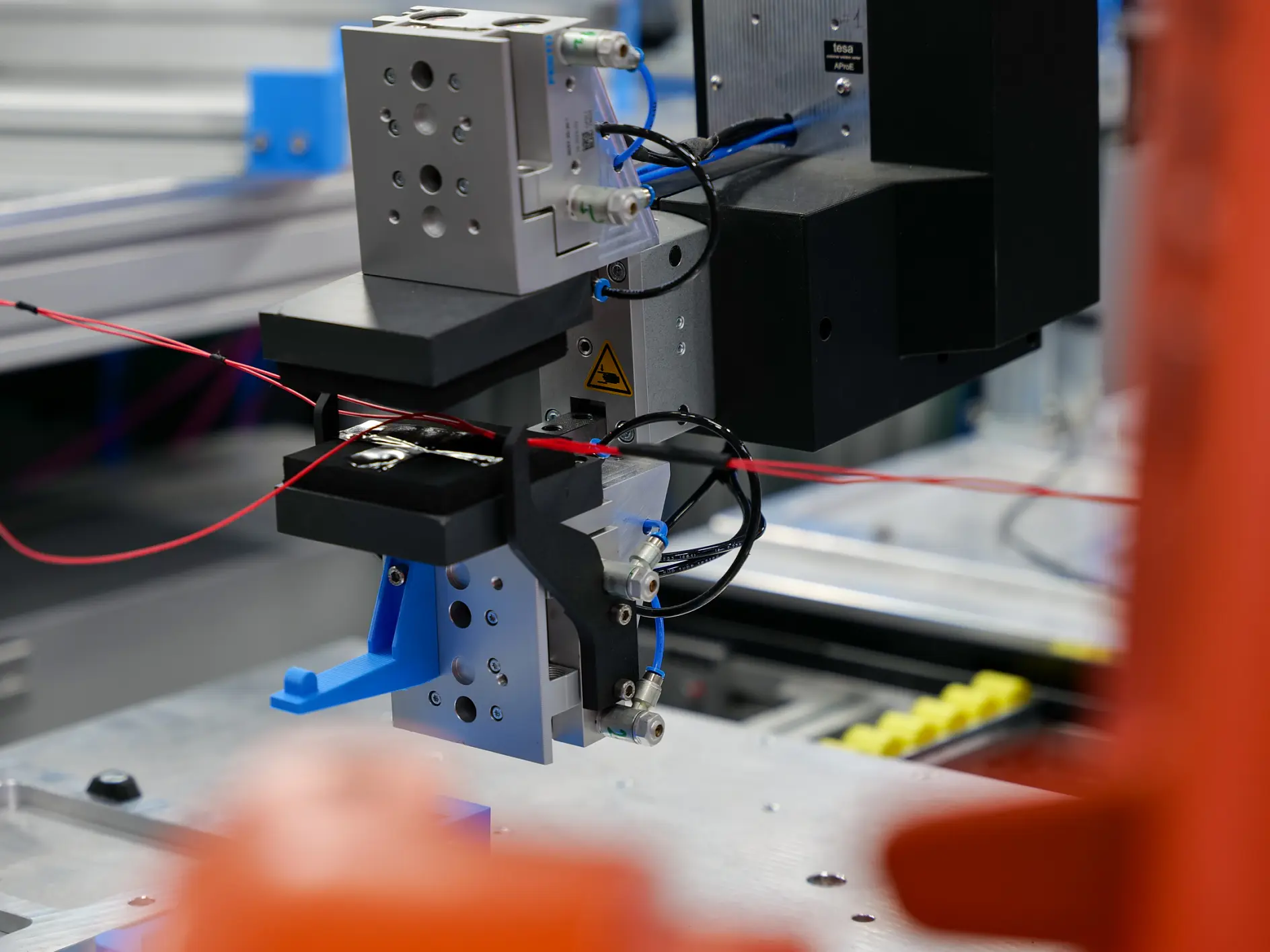 A close-up of an industrial robotic arm handling red wires and tesa tape on a manufacturing assembly line, with components visible. (This text has been generated by AI)