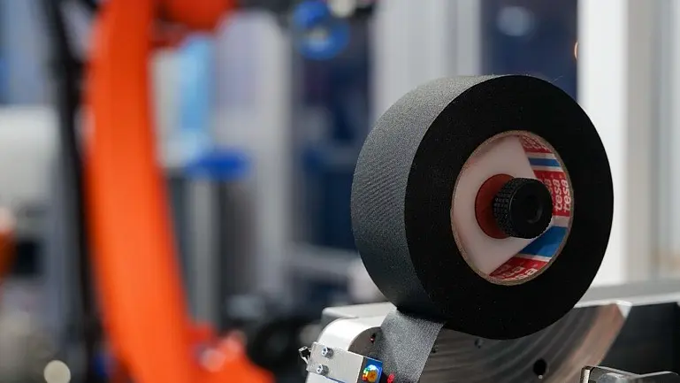 A close-up of a machine part with a black foam roller in the foreground and tesa tape, plus an orange robotic arm blurred behind. (This text has been generated by AI)
