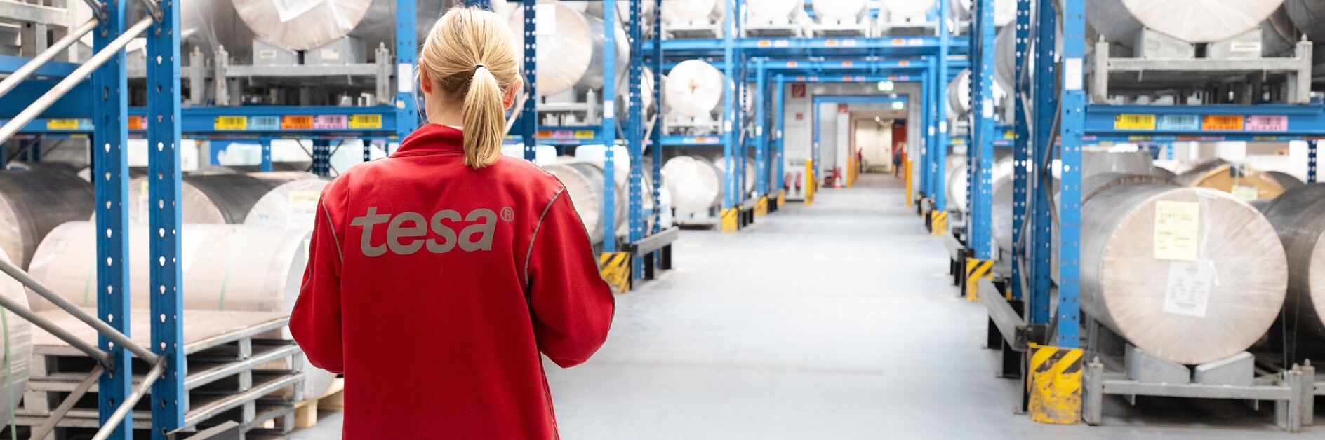 A person in a red jacket with tesa on the back stands in a warehouse aisle, surrounded by large rolls of tesa tape on blue shelves.