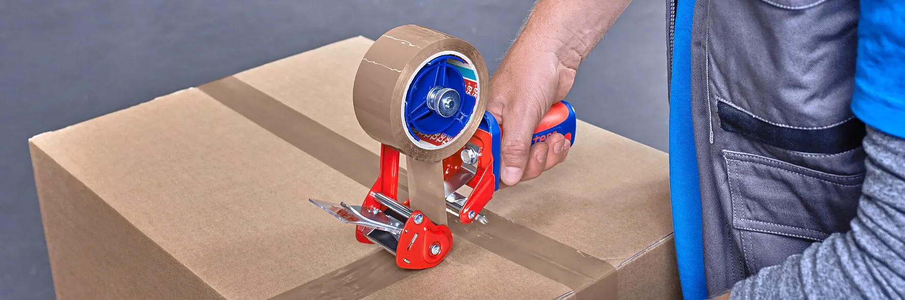 man sealing cardboard box with red tesa tape dispenser in warehouse filled with stacked boxes and shelves