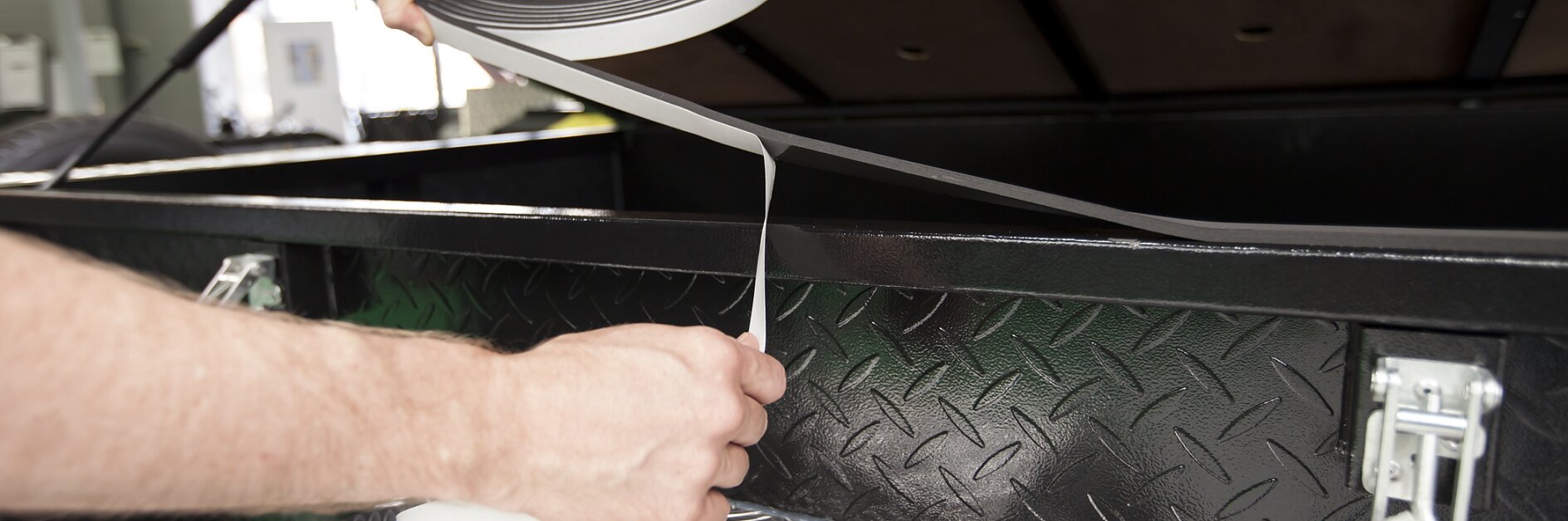 Hands are applying tesa tape to the edge of a black metal toolbox with a diamond plate pattern. The toolbox lid is open, and the interior is partly visible. A lock mechanism with a red latch is attached to the side.