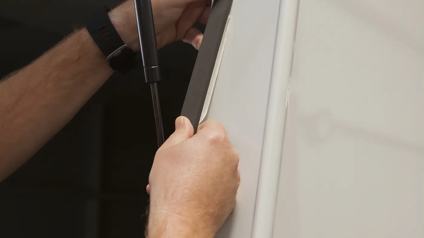 A person is applying a black tesa tape strip to the edge of a white panel. The panel is supported by a gas strut, and the hands are firmly pressing the tesa tape strip into place. The background is out of focus, showing a metallic structure.