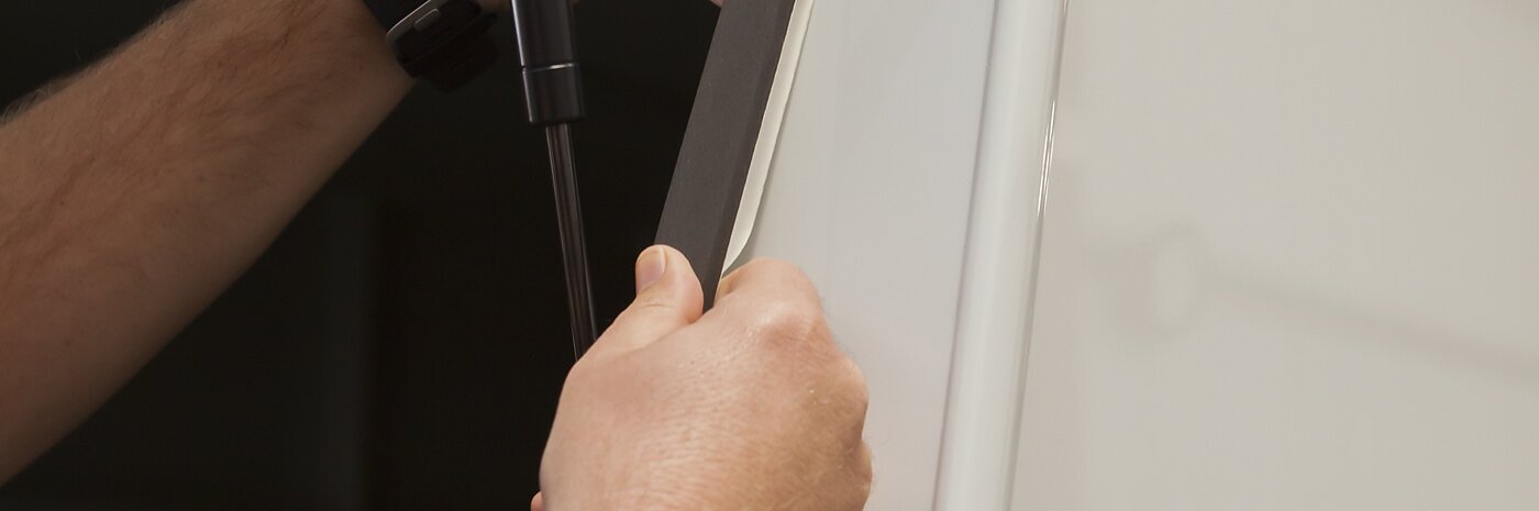 A person is applying a black tesa tape strip to the edge of a white panel. The panel is supported by a gas strut, and the hands are firmly pressing the tesa tape strip into place. The background is out of focus, showing a metallic structure.