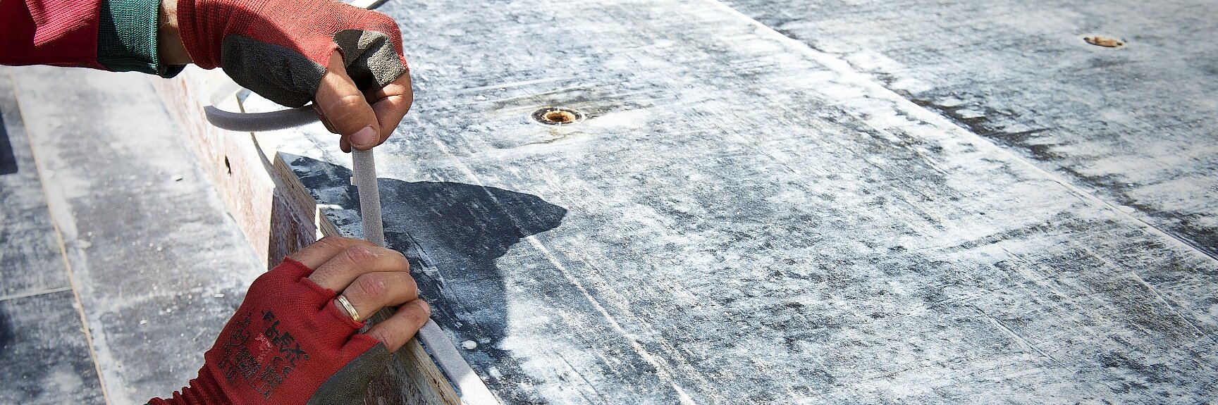 A person wearing red gloves is using a knife to cut a thick white cable on a concrete surface. The scene appears to be outdoors, with the surface showing signs of wear and weathering. The persons sleeves are pink and green. (This text has been generated by AI)