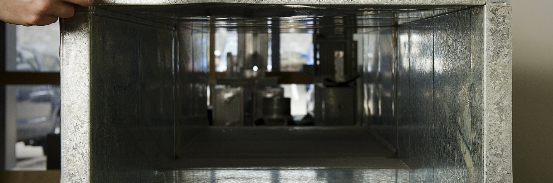 A person is measuring the edge of a metal air duct with a tesa tape measure. The duct is positioned horizontally, and the background shows blurred elements of an interior space. (This text has been generated by AI)