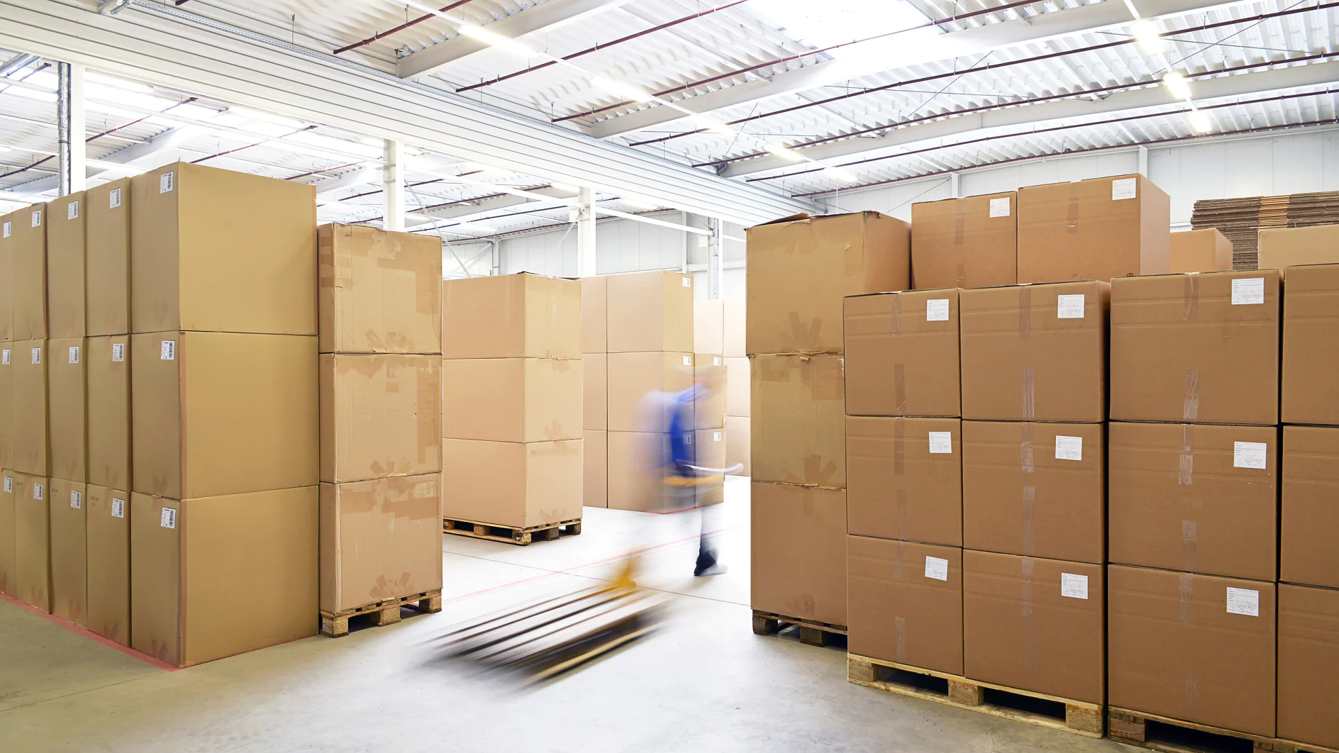 A large warehouse with stacks of cardboard boxes on wooden pallets. A person using a hand pallet truck is moving between the rows. The space is well-lit with natural light coming through the ceiling. The floor is concrete, and tesa tape is used throughout for securing packages and ensuring everything stays in place during transport. (This text has been generated by AI)