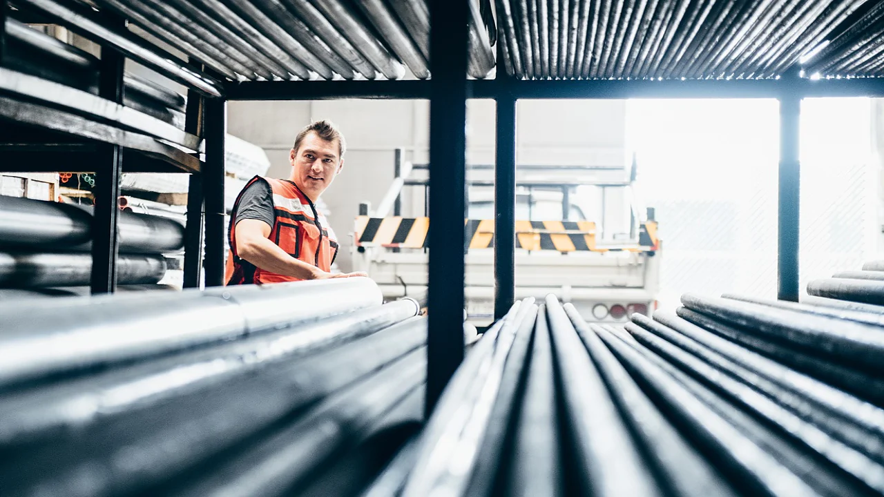 tesa растворы клейкой ленты металлургическая промышленность A person wearing an orange safety vest stands in a warehouse filled with metal pipes. They are looking to the side, surrounded by the stacked pipes on metal racks, with a truck partially visible in the background. (This text has been generated by AI)