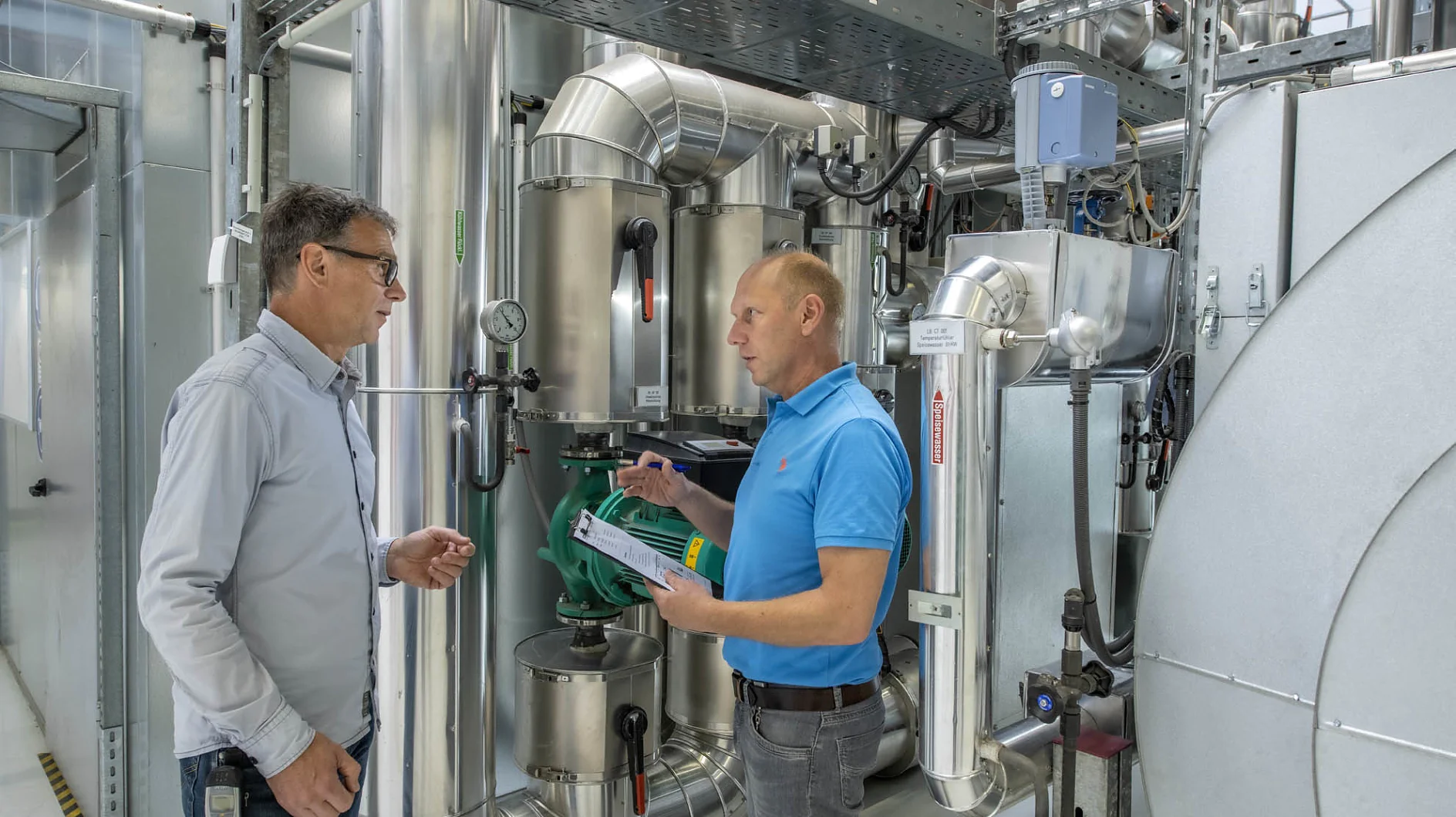 Two men are standing in an industrial facility filled with metallic pipes and machinery. One man, dressed in a blue shirt, holds a document while the other, wearing a white shirt, listens attentively. They seem to be discussing the equipment around them and considering the use of tesa tape for some applications. (This text has been generated by AI)