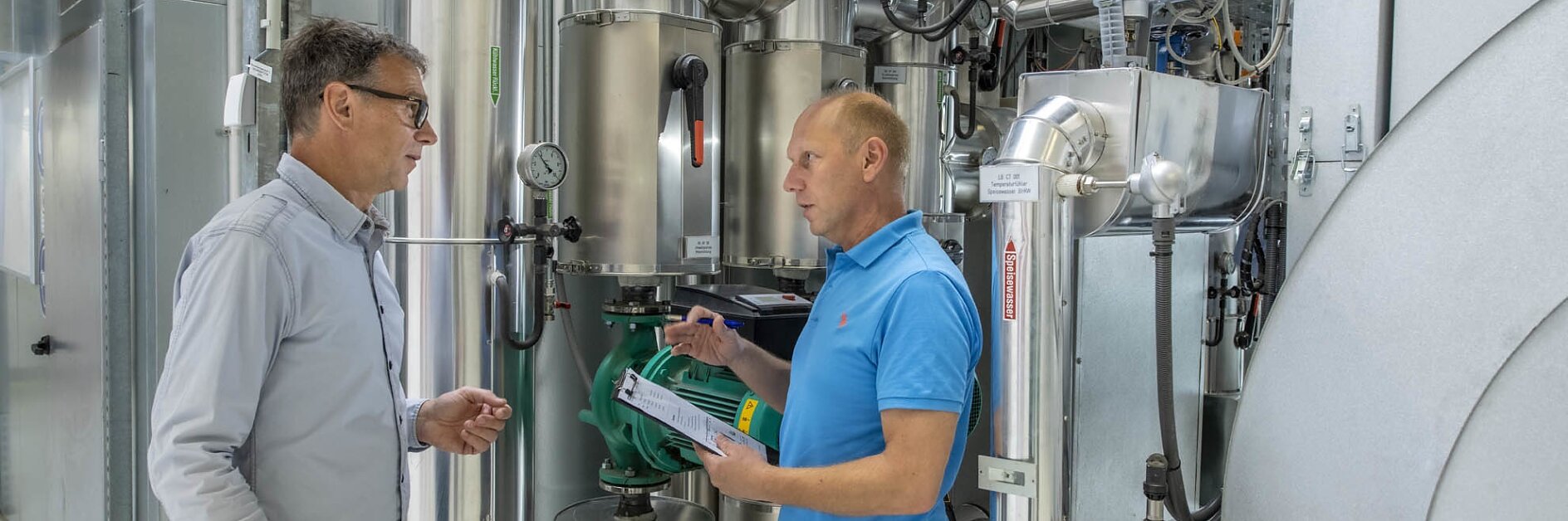 Two men are standing in an industrial facility filled with metallic pipes and machinery. One man, dressed in a blue shirt, holds a document while the other, wearing a white shirt, listens attentively. They seem to be discussing the equipment around them and considering the use of tesa tape for some applications. (This text has been generated by AI)