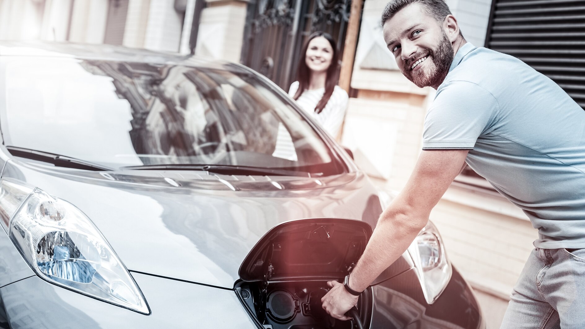 E-Mobility Key Visual Easy and ecological transportation. Selective focus on a happy millennial guy grinning broadly while holding a charging nozzle and enjoying the process of his eco car supplying energy outdoors.