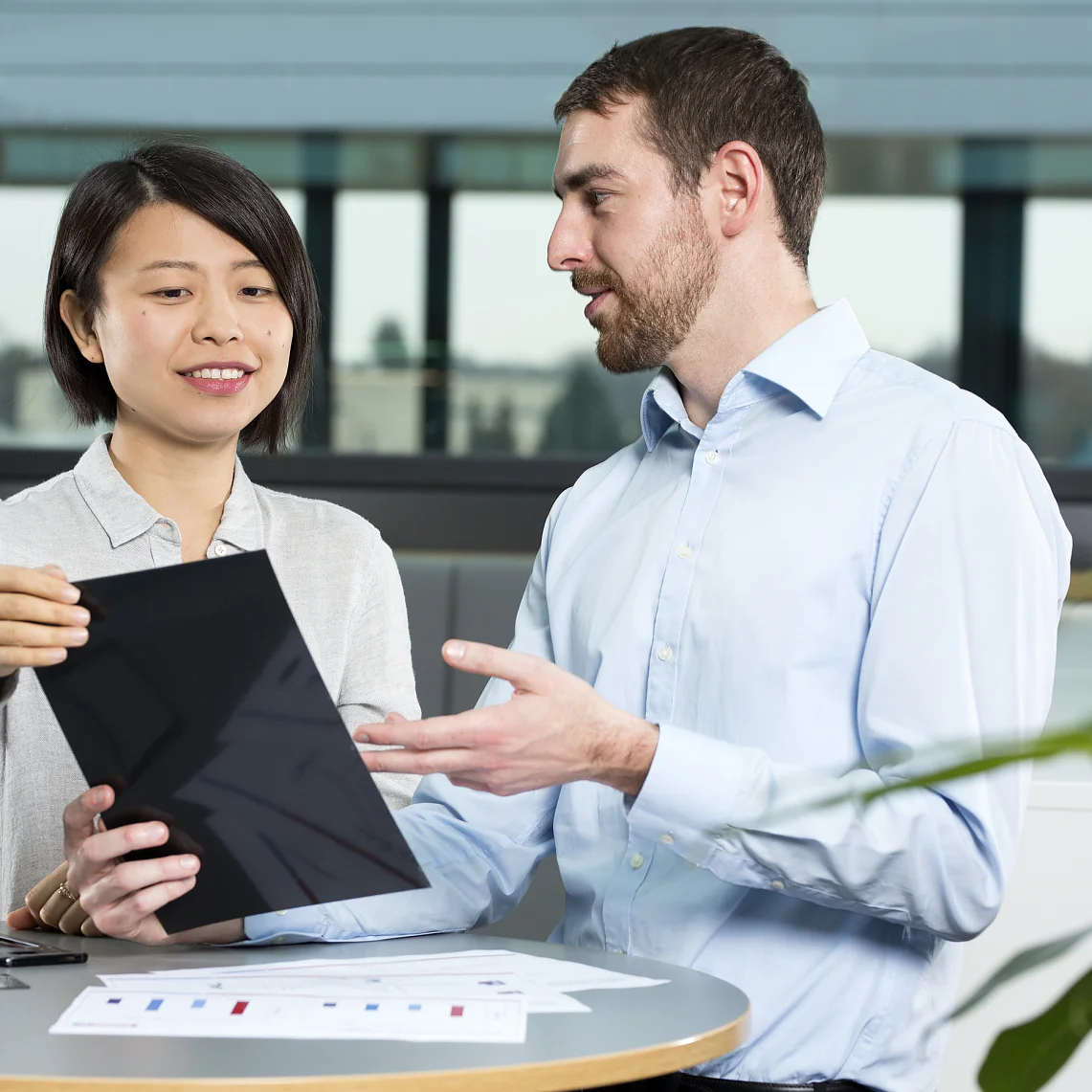 Two people are discussing something on a tablet in an office setting. The woman is holding the tablet, while the man gestures toward it. A document with graphs is on the table secured with tesa tape. They are seated near a window with a view outside. (This text has been generated by AI)