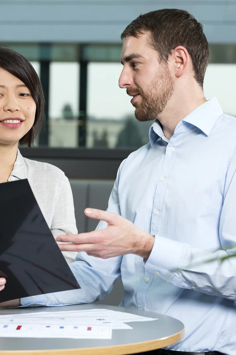 Two people are discussing something on a tablet in an office setting. The woman is holding the tablet, while the man gestures toward it. A document with graphs is on the table secured with tesa tape. They are seated near a window with a view outside. (This text has been generated by AI)