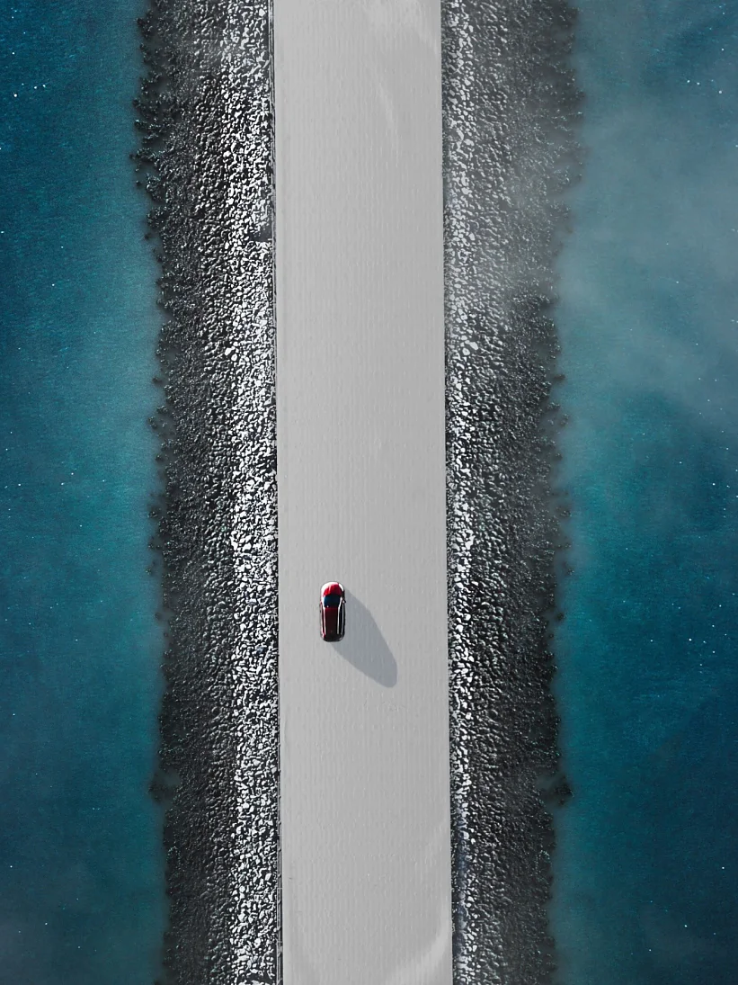 Aerial view of a red car driving on a narrow, straight road flanked by blue water on both sides. The road is elevated, with rocky edges lining the sides. The water appears calm with a slight mist or fog hovering above it. (This text has been generated by AI)