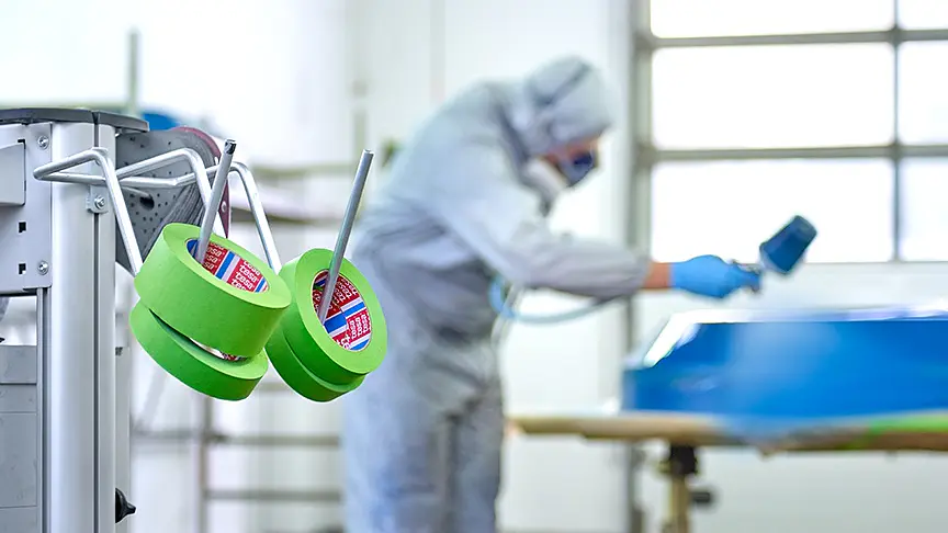 A person wearing protective gear and a respirator is spray painting a blue object in a workshop. Rolls of green tesa tape hang on a stand in the foreground. The workshop has a large window in the background.