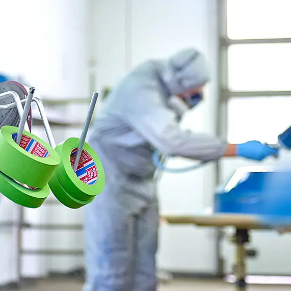 A person wearing protective gear and a respirator is spray painting a blue object in a workshop. Rolls of green tesa tape hang on a stand in the foreground. The workshop has a large window in the background. (This text has been generated by AI)