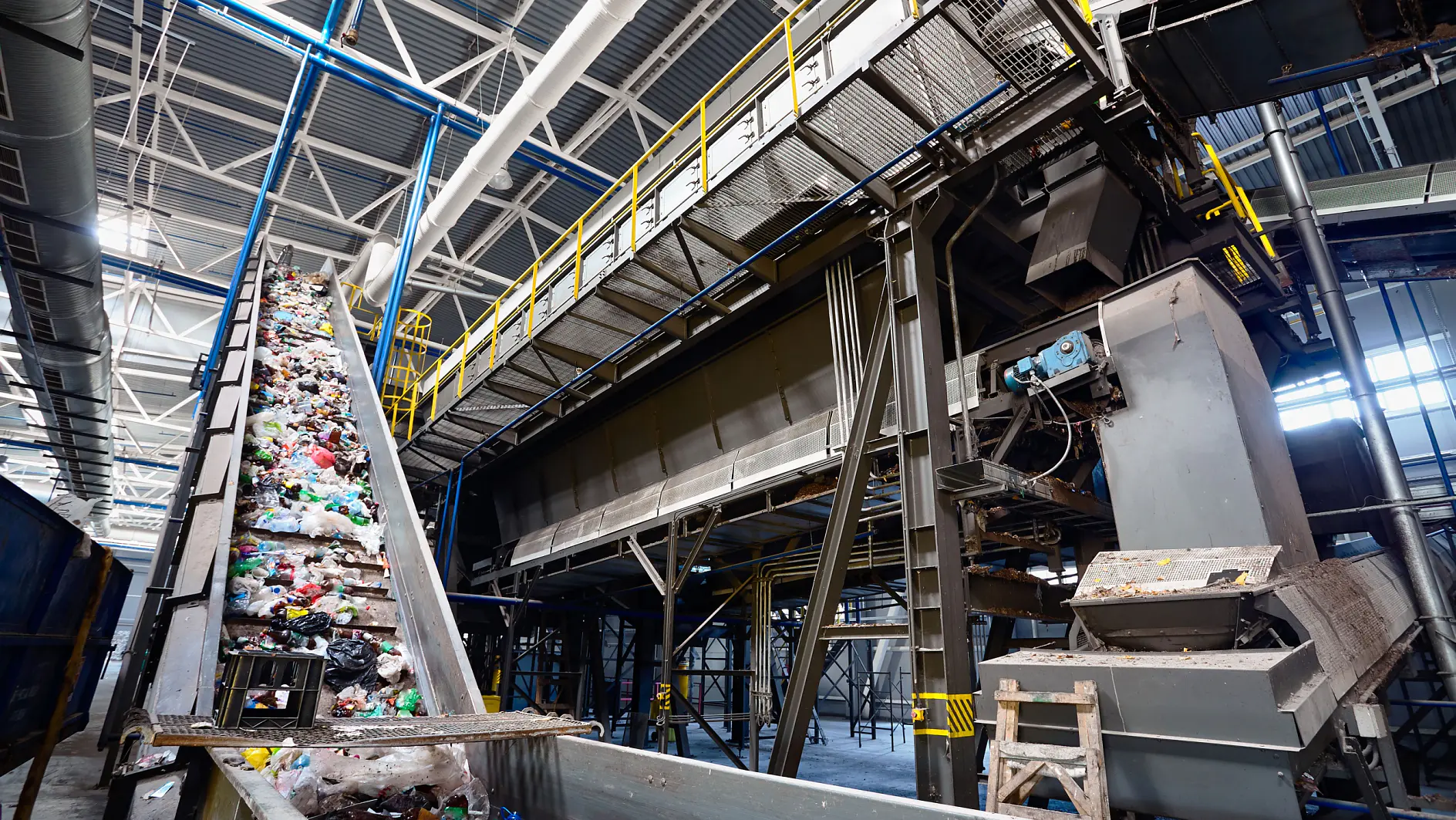 Conveyor belt carrying assorted waste materials in a large industrial recycling facility. Overhead metal structures and machinery are visible, with a high ceiling and hanging lights providing illumination.