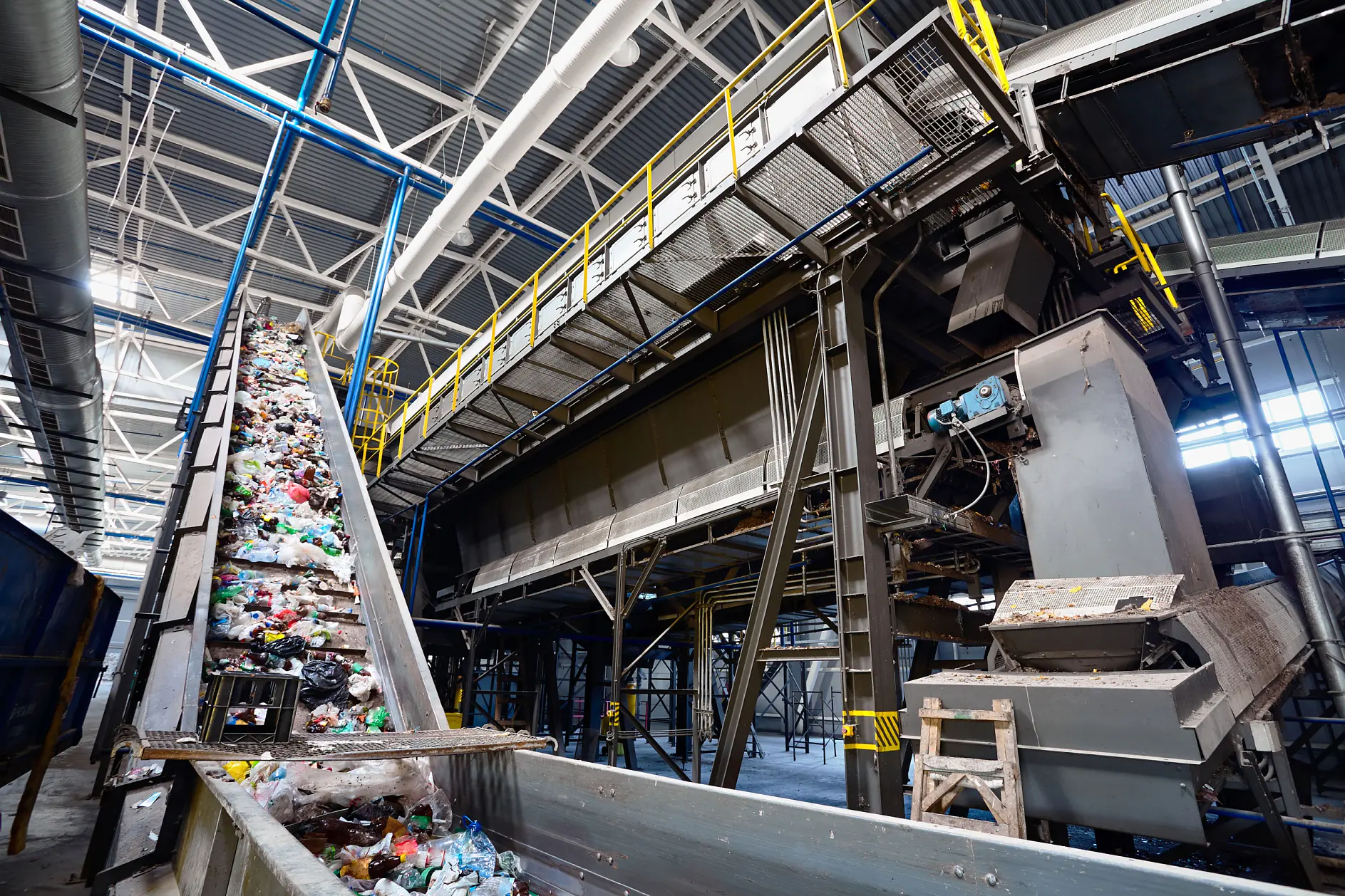 Conveyor belt carrying assorted waste materials in a large industrial recycling facility. Overhead metal structures and machinery are visible, with a high ceiling and hanging lights providing illumination.