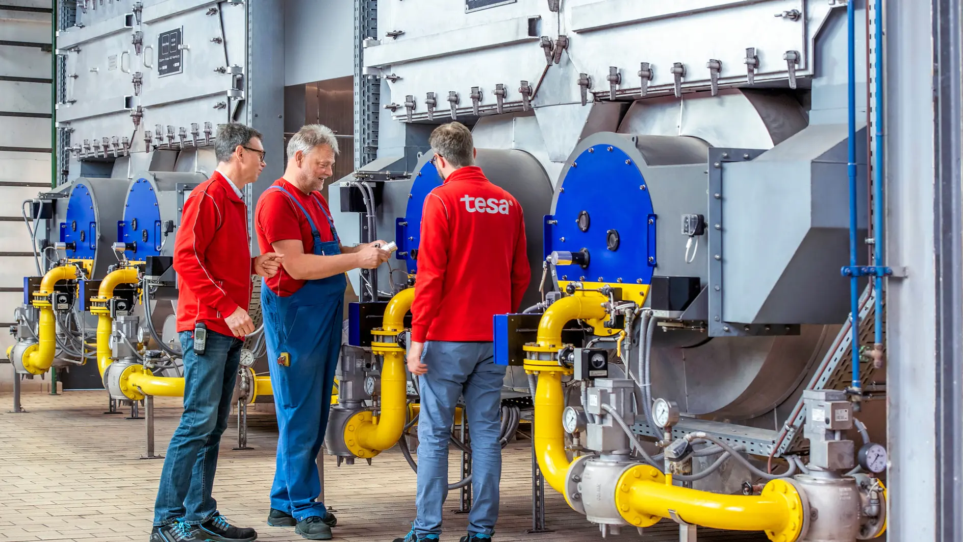 Three men stand in front of large industrial machinery with blue and silver components. They are wearing red shirts and are having a discussion. Yellow pipes are connected to the machines, and the floor is paved with tiles. (This text has been generated by AI)