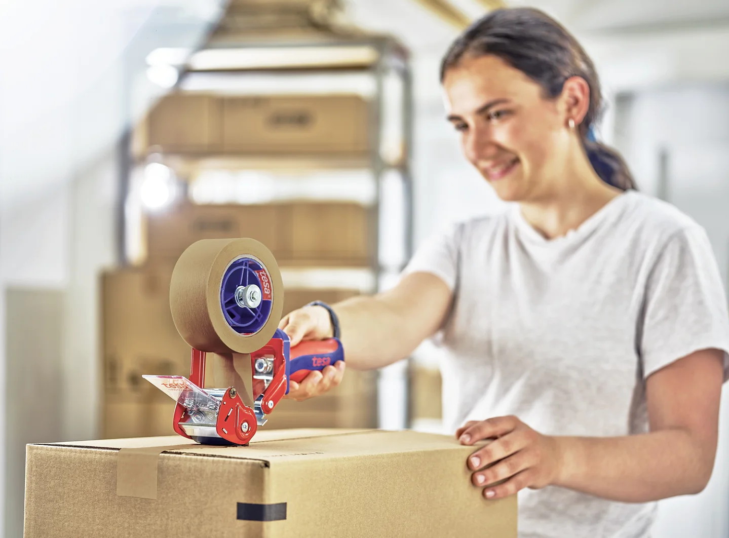 A person in a white shirt uses a handheld tesa tape dispenser to seal a cardboard box. They are in a room with several stacked boxes in the background and appear to be smiling. (This text has been generated by AI)