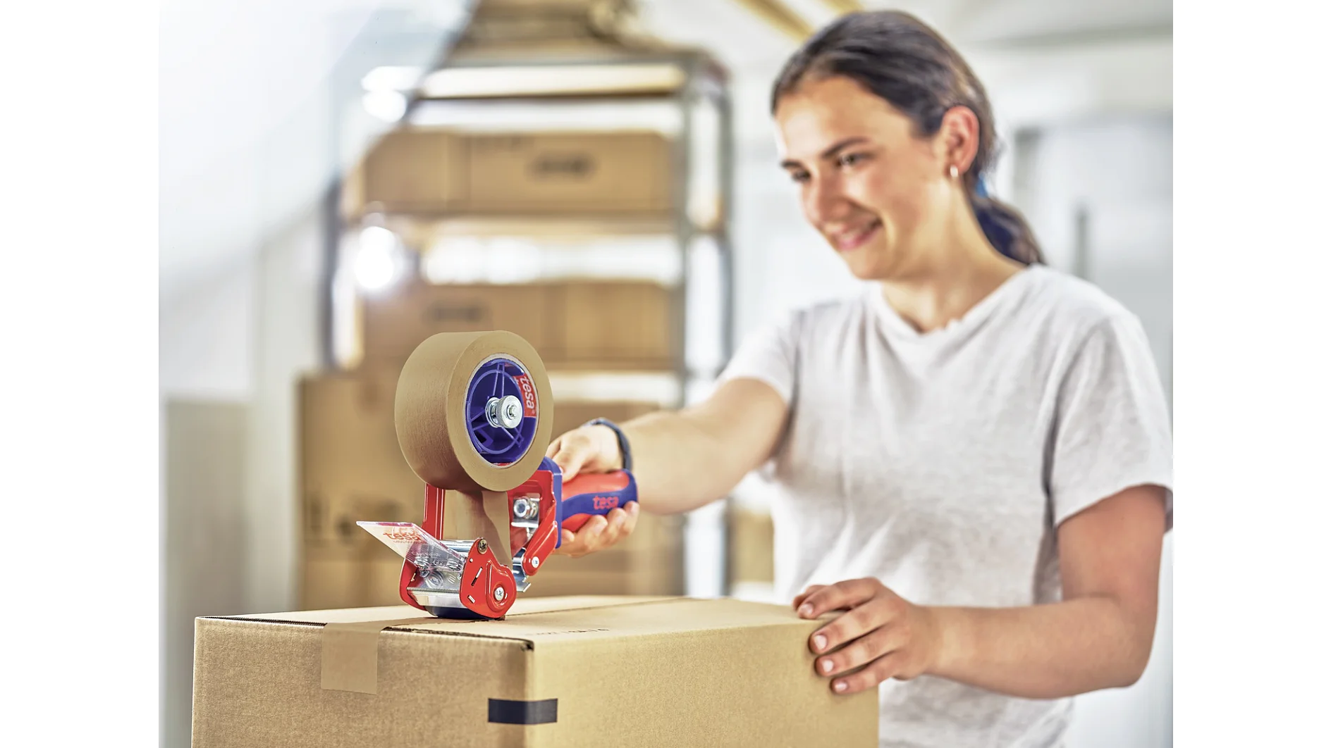 A person in a white shirt uses a handheld tesa tape dispenser to seal a cardboard box. They are in a room with several stacked boxes in the background and appear to be smiling. (This text has been generated by AI)