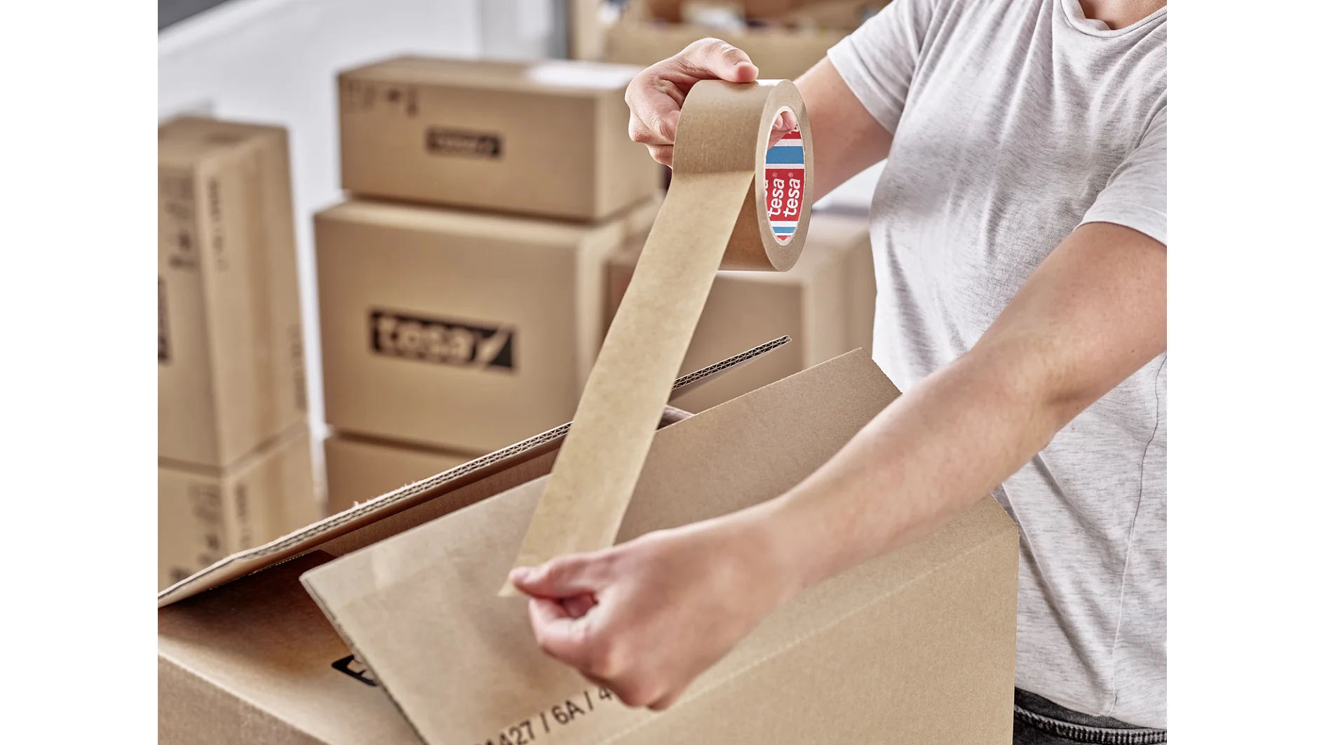 A person in a white shirt is sealing a cardboard box with brown packing tesa tape. In the background, several other cardboard boxes are stacked. The scene is indoors, suggesting a packing or moving process. (This text has been generated by AI)