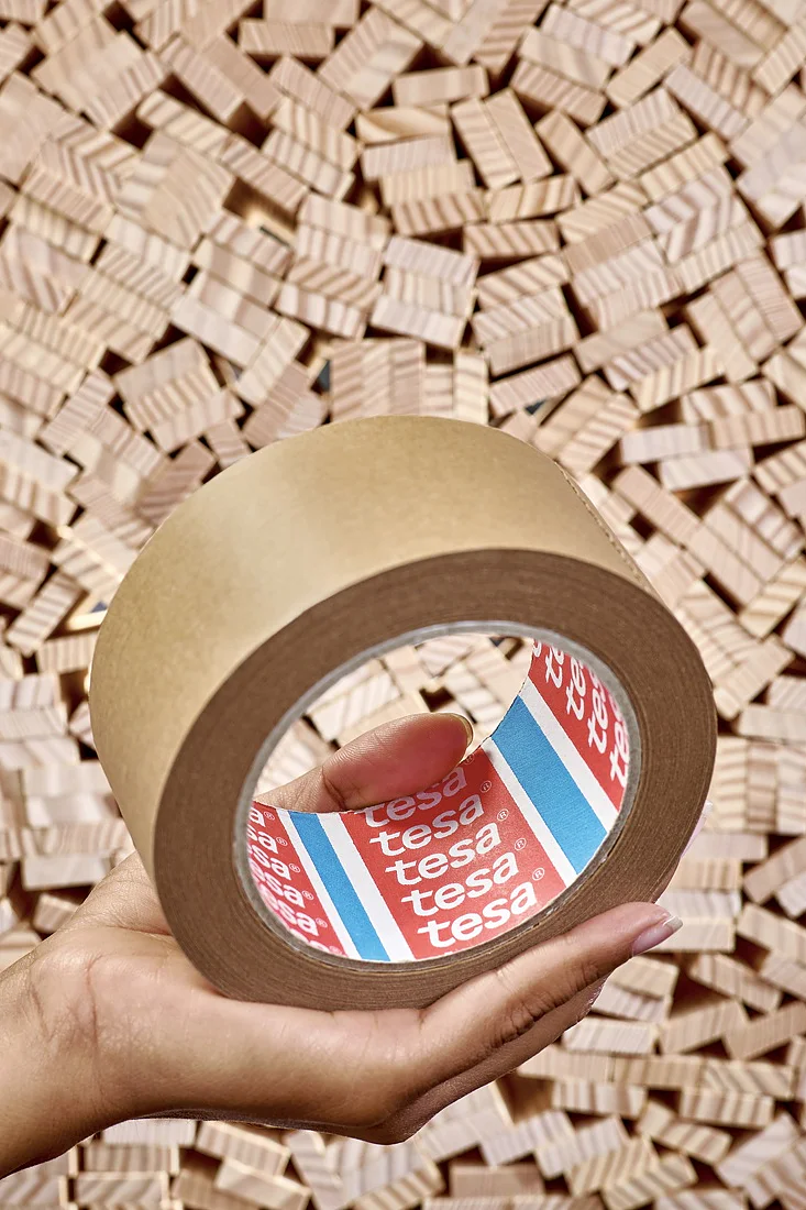 A hand holds a roll of brown tesa tape labeled tesa in red and white. The background consists of numerous small, wooden blocks stacked tightly together. (This text has been generated by AI)