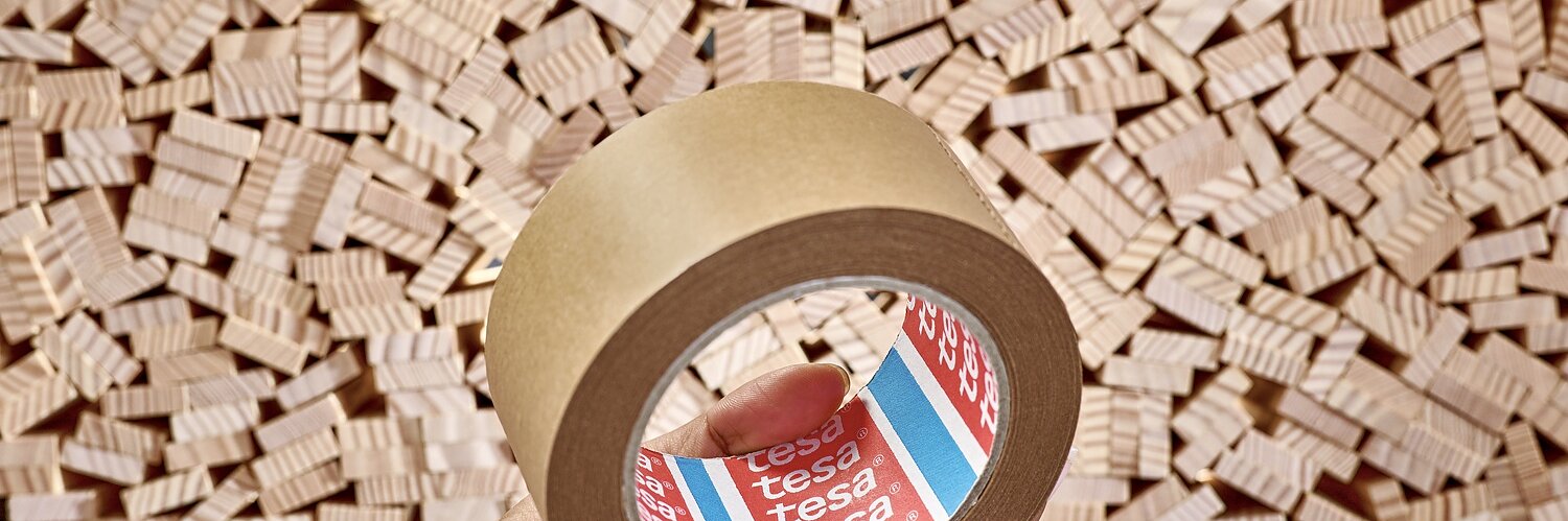 A hand holds a roll of brown tesa tape labeled tesa in red and white. The background consists of numerous small, wooden blocks stacked tightly together. (This text has been generated by AI)