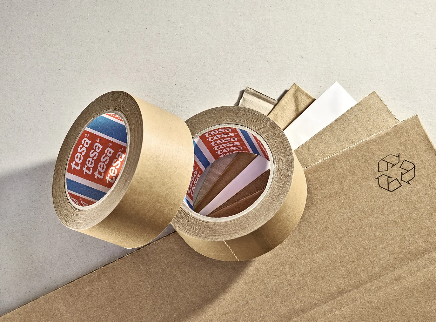 Two rolls of brown adhesive tesa tape are placed on a surface along with several sheets of cardboard. One roll is upright, while the other is lying down, showing the white inner side with the brand name visible. A recycling symbol is printed nearby. (This text has been generated by AI)