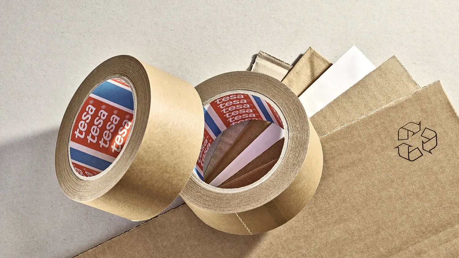 4513-superior-paper-packaging-tape-mp (3) Two rolls of brown adhesive tesa tape are placed on a surface along with several sheets of cardboard. One roll is upright, while the other is lying down, showing the white inner side with the brand name visible. A recycling symbol is printed nearby. (This text has been generated by AI)