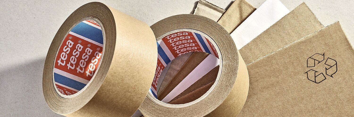 Two rolls of brown adhesive tesa tape are placed on a surface along with several sheets of cardboard. One roll is upright, while the other is lying down, showing the white inner side with the brand name visible. A recycling symbol is printed nearby. (This text has been generated by AI)