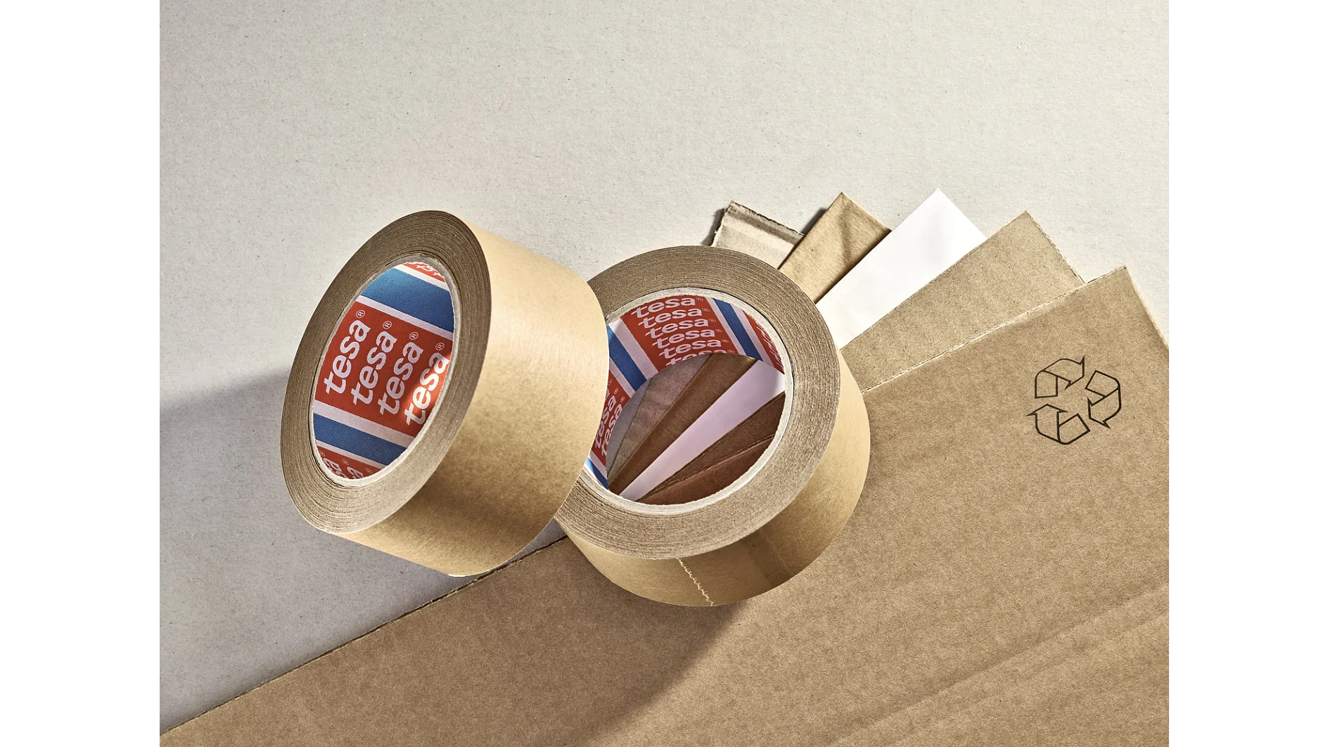 Two rolls of brown adhesive tesa tape are placed on a surface along with several sheets of cardboard. One roll is upright, while the other is lying down, showing the white inner side with the brand name visible. A recycling symbol is printed nearby. (This text has been generated by AI)