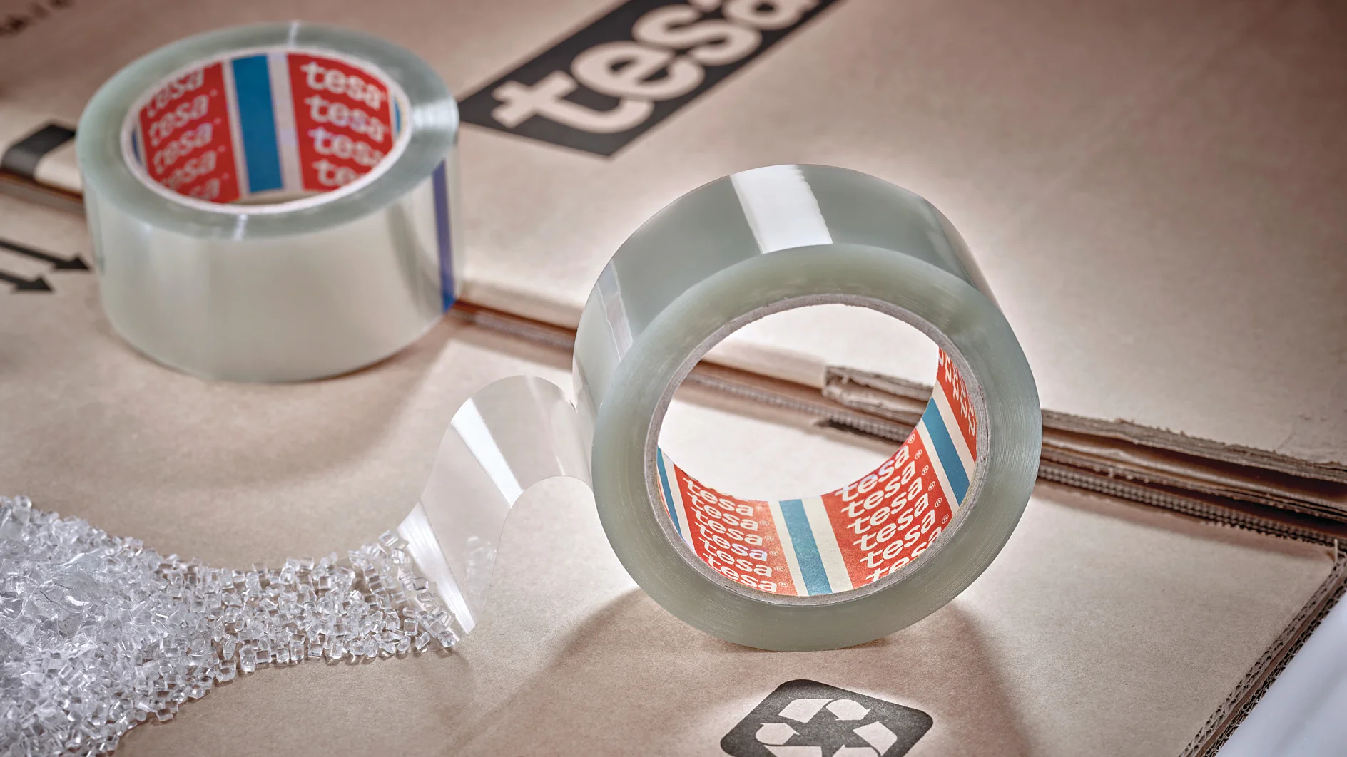 A roll of clear packing tesa tape with the brand name visible sits on a cardboard surface. A strip of tesa tape extends outward. Another roll is partially visible, and bubble wrap is nearby. The cardboard has a recycling symbol printed on it. (This text has been generated by AI)