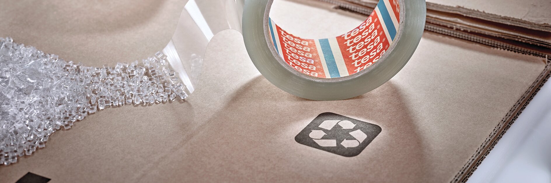 A roll of clear packing tesa tape with the brand name visible sits on a cardboard surface. A strip of tesa tape extends outward. Another roll is partially visible, and bubble wrap is nearby. The cardboard has a recycling symbol printed on it. (This text has been generated by AI)