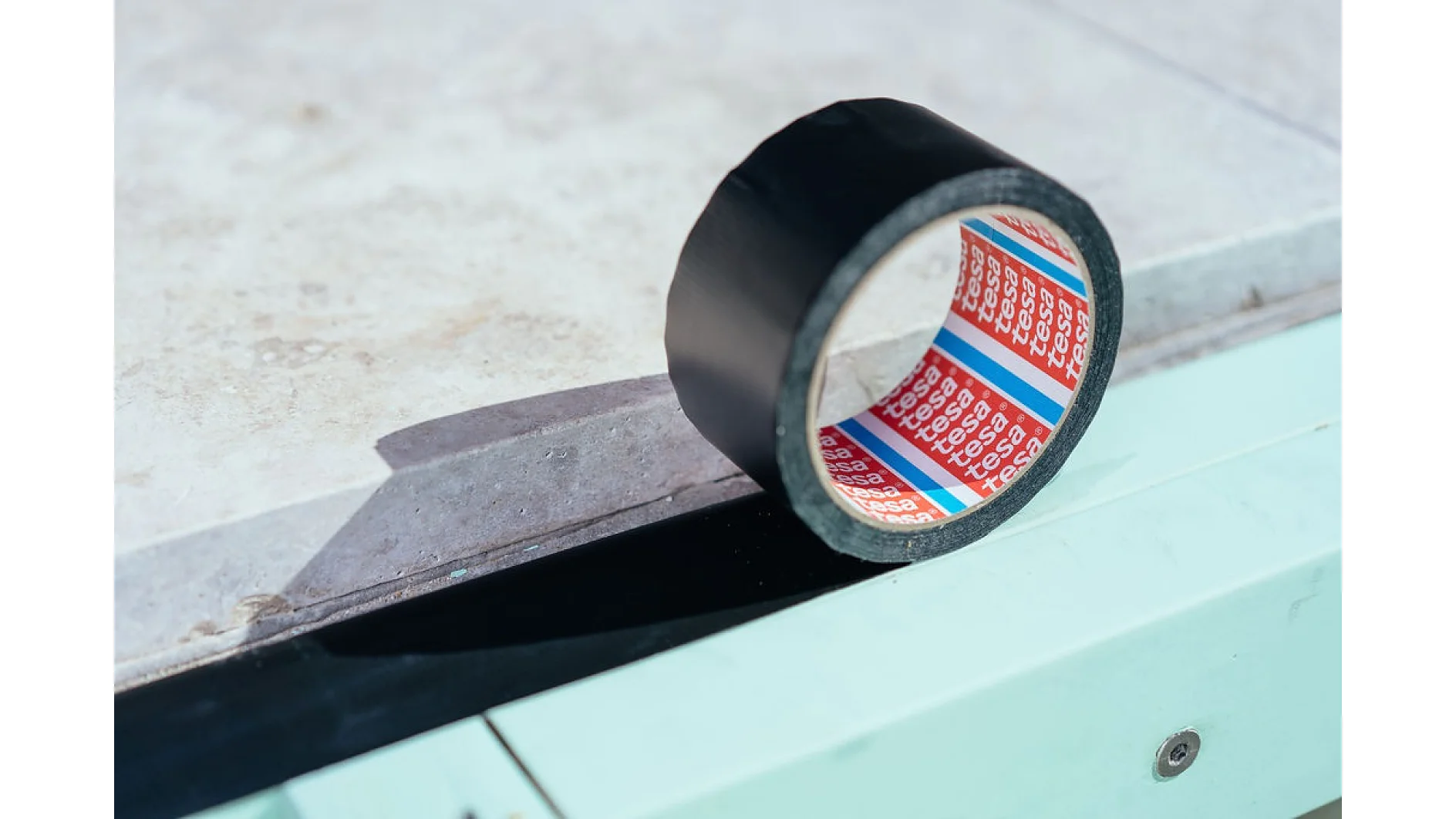 A roll of black tesa tape sits on a light-colored surface. The inner side of the tesa tape displays a red and blue pattern with white text. A strip of the tesa tape is partially unrolled and adhered to the surface. (This text has been generated by AI)