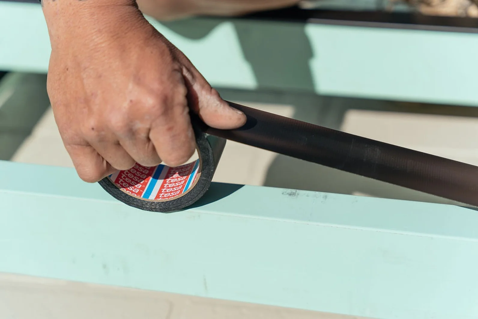 A hand is holding a roll of black tesa tape, preparing to apply it to a light green surface. The tesa tape shows a red and white label inside the roll. The scene appears to be outdoors in natural light. (This text has been generated by AI)