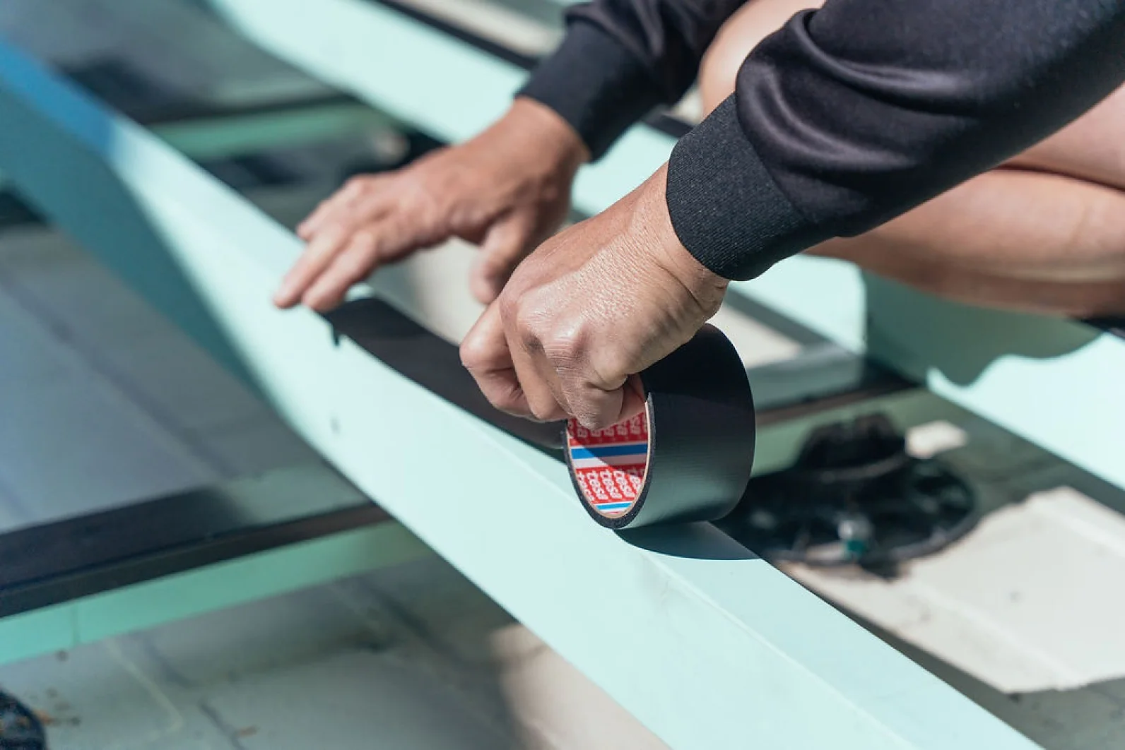 A person wearing a black sleeve is applying black tesa tape to a light blue metal beam. The setting appears to be outdoors, and the focus is on the hands holding the tesa tape, indicating construction or repair work in progress. (This text has been generated by AI)
