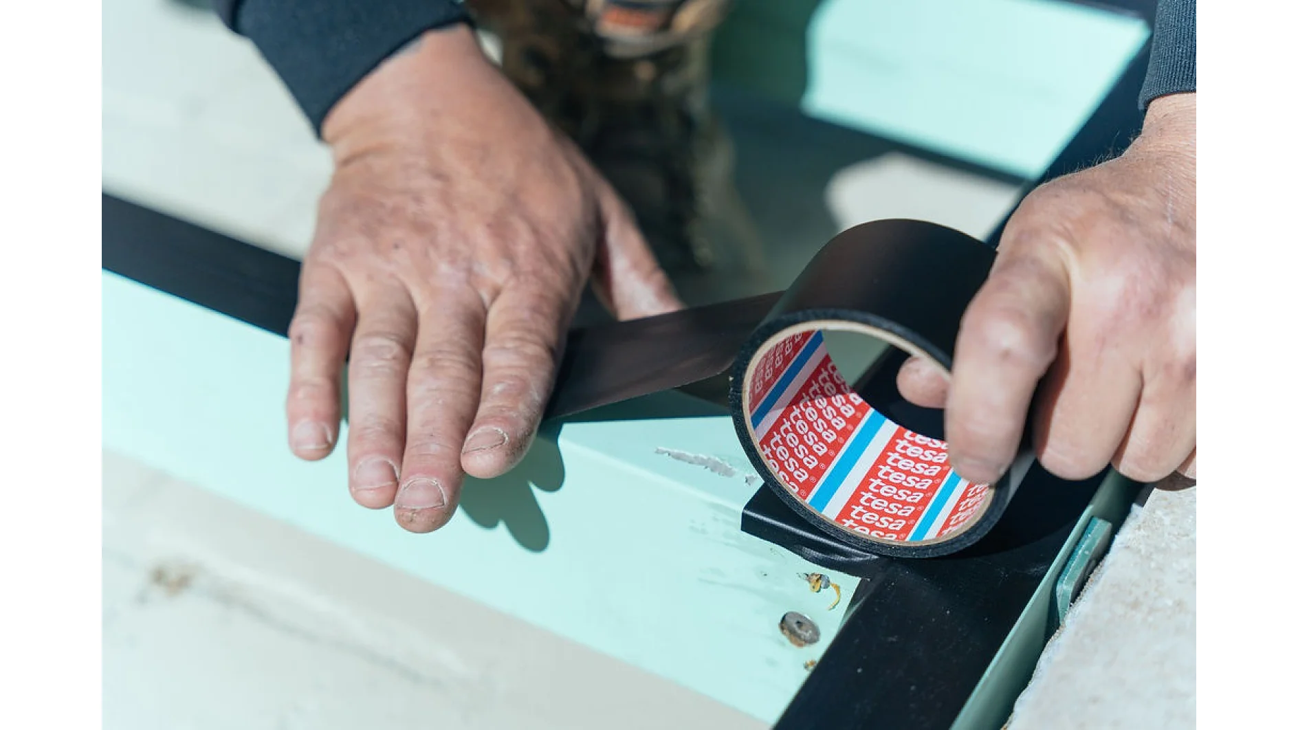A person is applying black tesa tape to a light blue surface with hands visible. The person is using the tesa tape directly from the roll, and a corner of the surface is partially taped. The background is out of focus. (This text has been generated by AI)