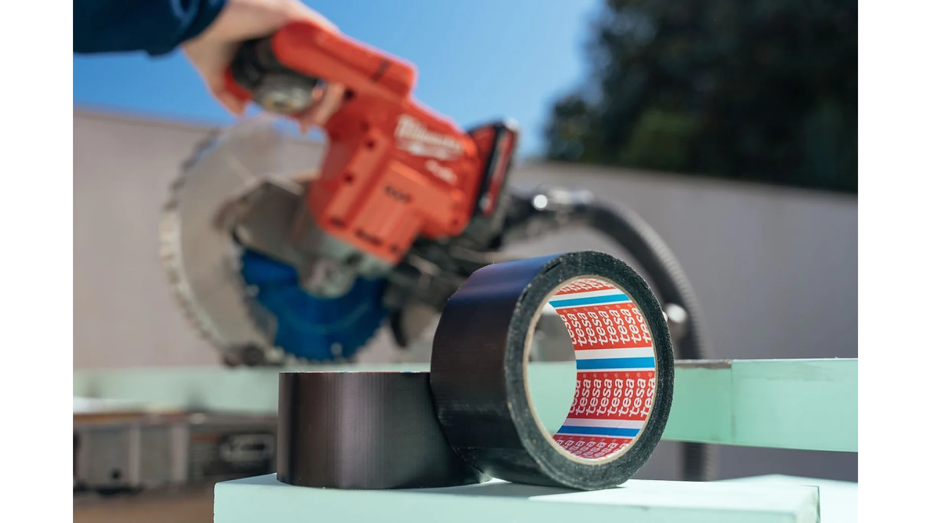 A roll of black tesa tape sits on a light green surface. In the blurred background, a person holds a red power tool, possibly a saw, with a silver blade. The scene appears to be outdoors with a wall and trees visible. (This text has been generated by AI)
