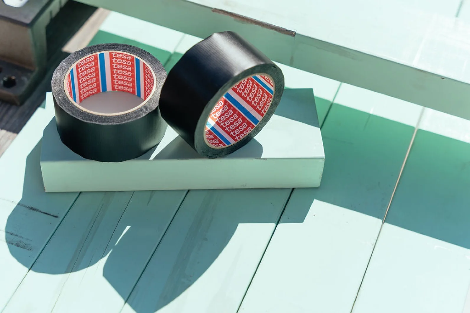 Two rolls of black tesa tape with red and white branded inner wraps rest on a light green wooden surface. They are positioned side by side in direct sunlight, casting shadows on the surface. (This text has been generated by AI)