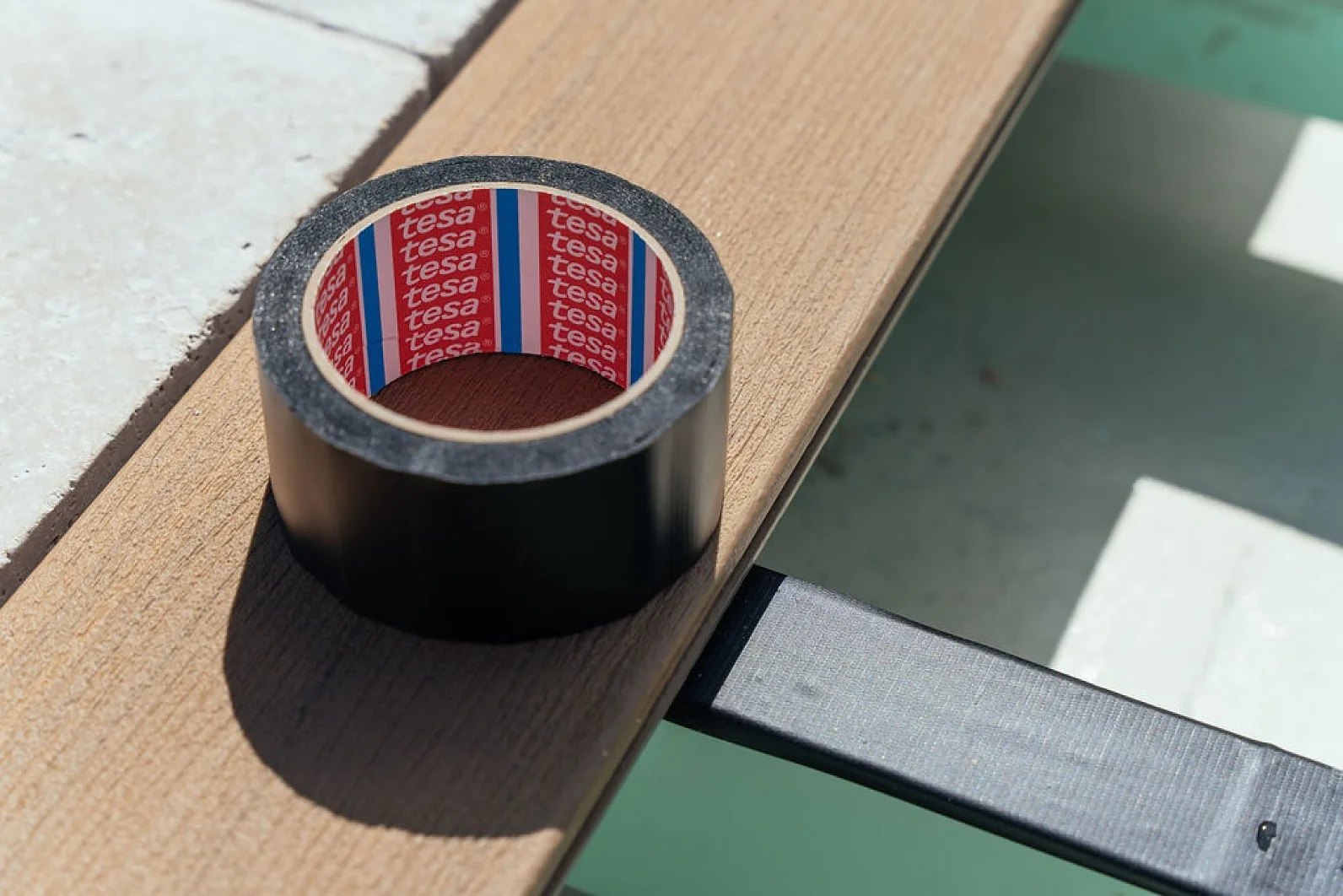 A roll of black tesa tape sits on a wooden board positioned next to a tiled surface. Sunlight casts shadows across the scene, creating a contrast between the light and dark areas. (This text has been generated by AI)