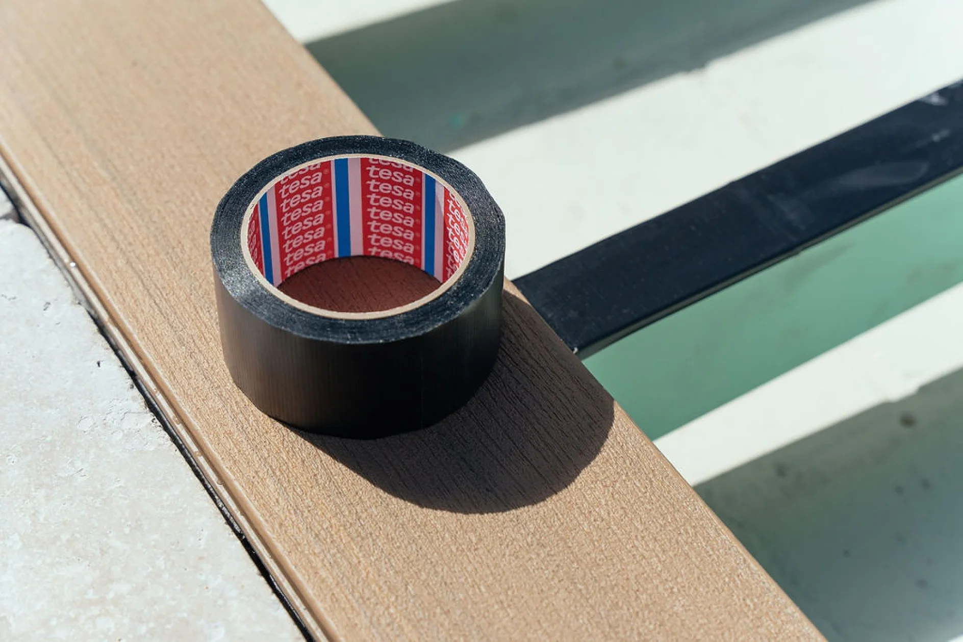 TESA_TAPE-36 A roll of black adhesive tesa tape is placed on a wooden plank. The interior of the tesa tape is red and features the brand name tesa in white. The plank is part of a structure with metal beams and a light background. (This text has been generated by AI)