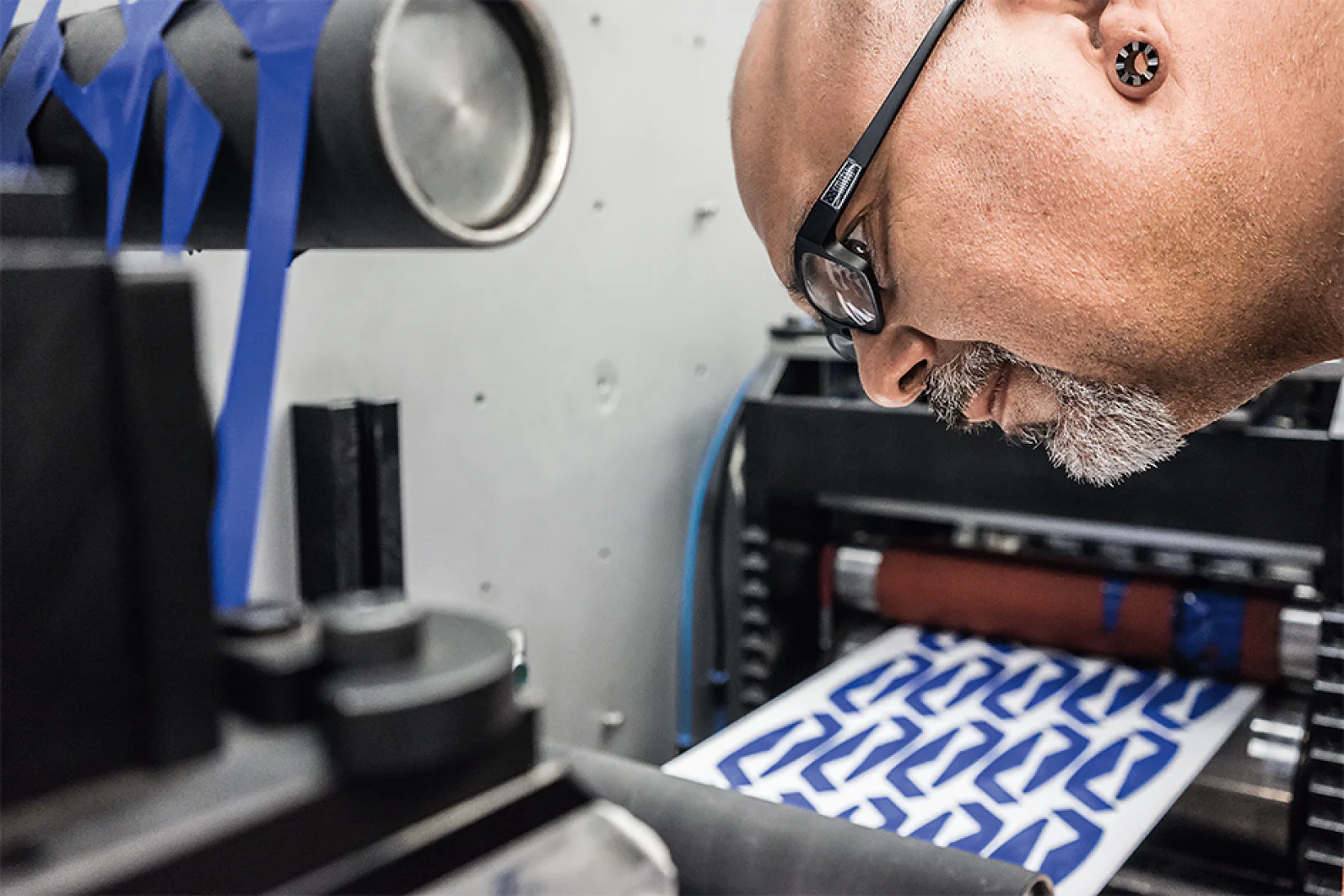 A person with glasses and earrings inspects a printing machine. Blue and white patterned paper emerges from the machine secured with tesa tape. The background is industrial, with visible machinery and rollers reinforced by tesa tape for stability. (This text has been generated by AI)