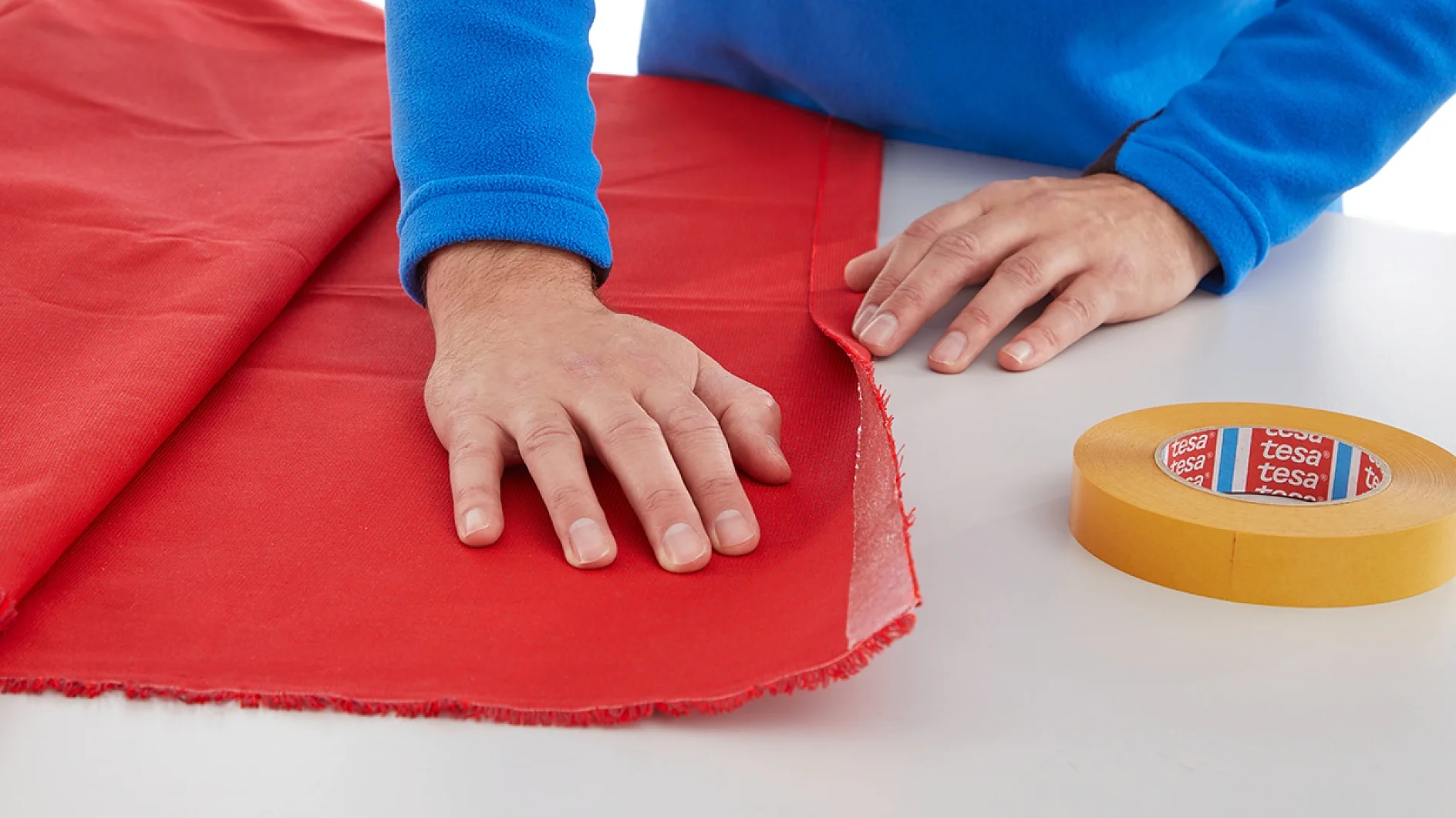 A person in a blue sweater smooths out a piece of red fabric on a white table. A roll of yellow tesa tape is visible to the right. (This text has been generated by AI)