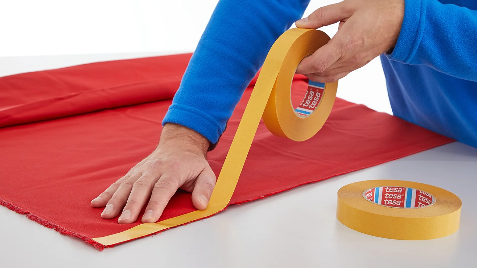 A person wearing a blue sweater is placing yellow tesa tape onto a piece of red fabric on a white table. Another roll of tesa tape is nearby. The person uses one hand to smooth the tesa tape while holding the roll with the other hand. (This text has been generated by AI)