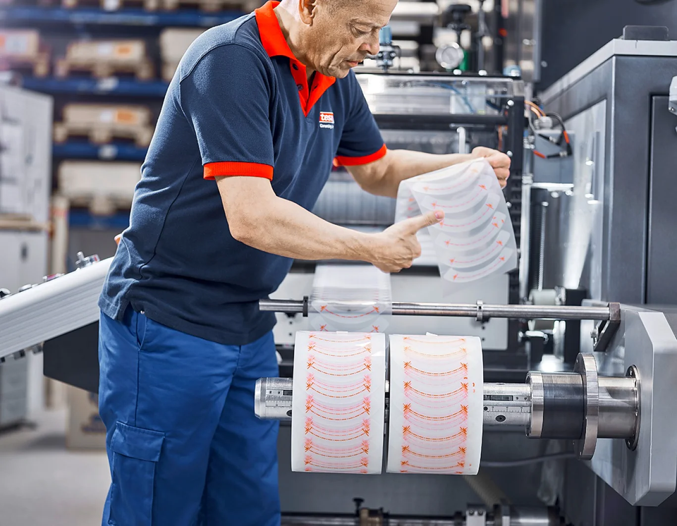 A worker in a navy and red uniform examines printed sheets on a machine. The machine is processing large rolls of material, and the worker is holding the sheets up for inspection. Shelving with items, including tesa tape, is visible in the background. (This text has been generated by AI)