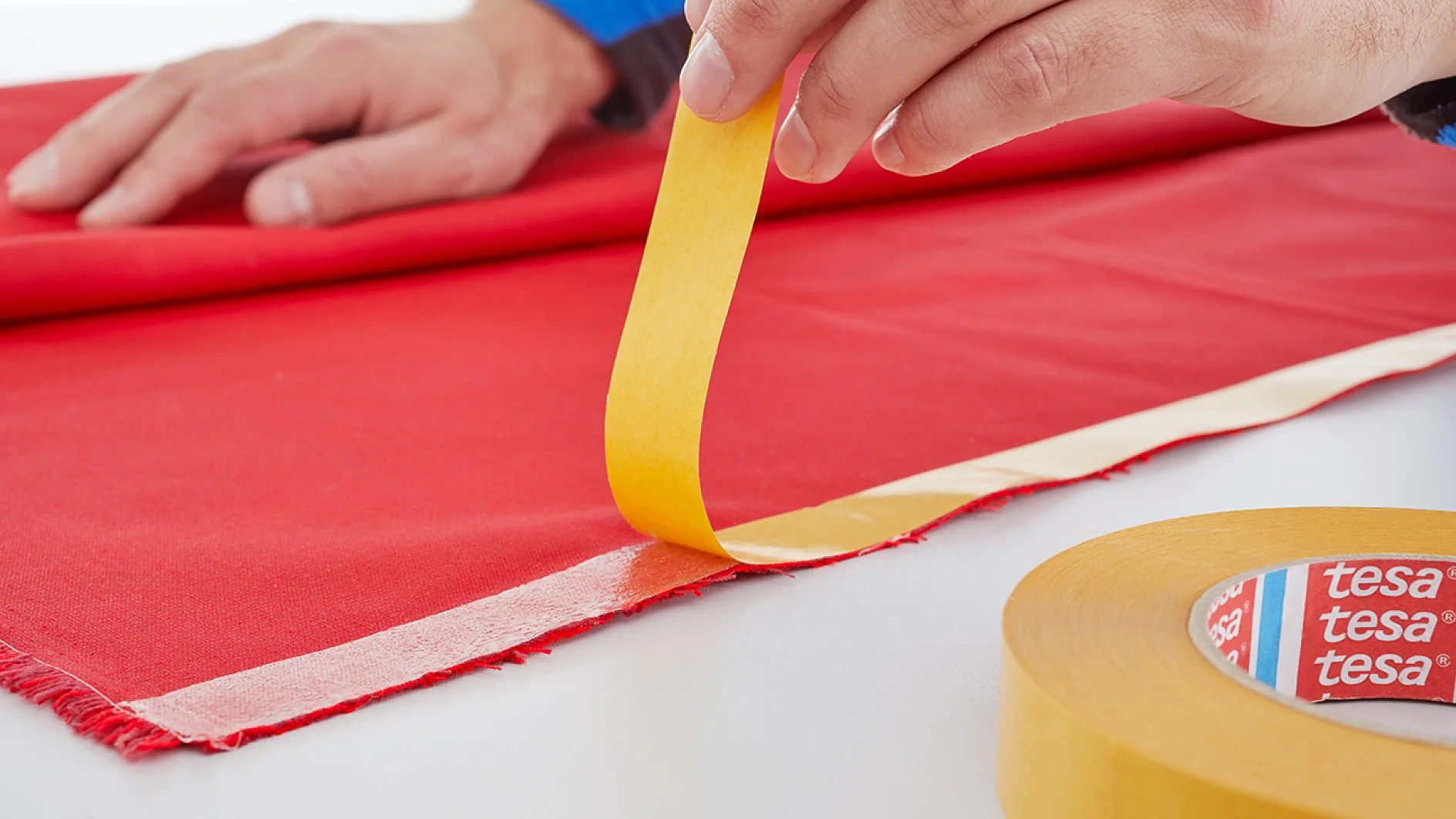 A person applies yellow tesa tape to the edge of a red fabric laid on a white surface. A roll of the same tesa tape is visible nearby. The brand name tesa is printed in red on the inner core of the roll. (This text has been generated by AI)