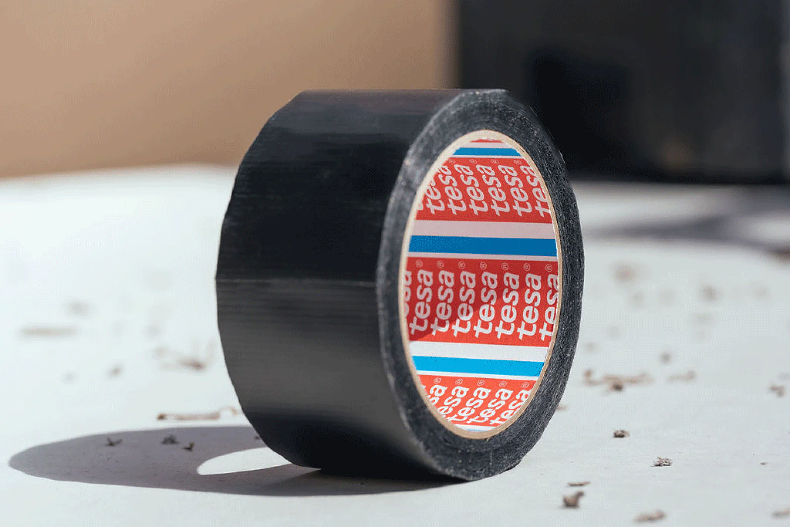 A roll of black adhesive tesa tape sits on a surface with debris. The inner part of the roll has red, blue, and white branding. The background is softly blurred, casting a subtle shadow from the tesa tape. (This text has been generated by AI)